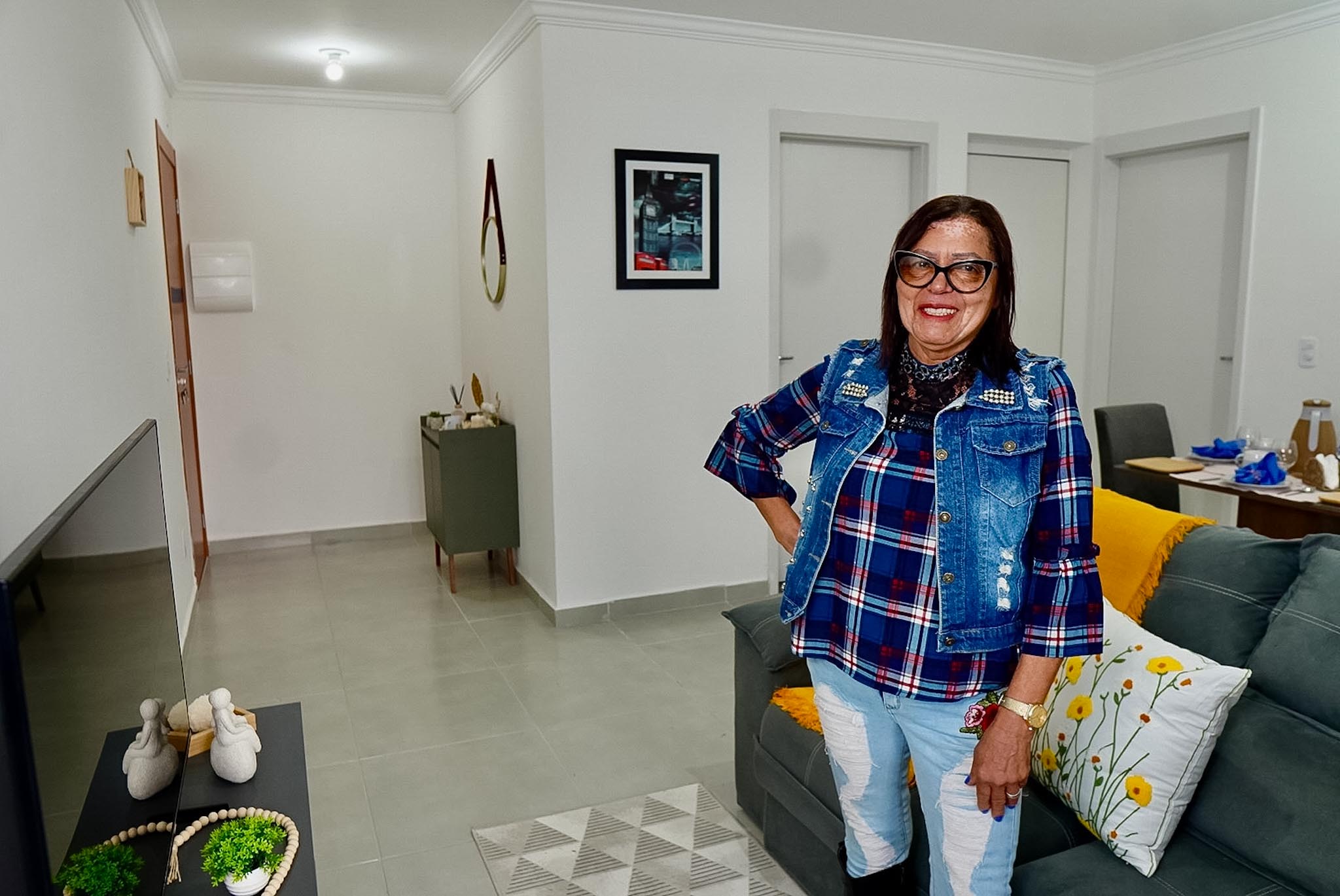 Mulher sorridente em uma sala de estar clara e organizada, em pé ao lado de um sofá cinza com almofadas decorativas. O ambiente possui paredes brancas, piso claro e decoração simples.