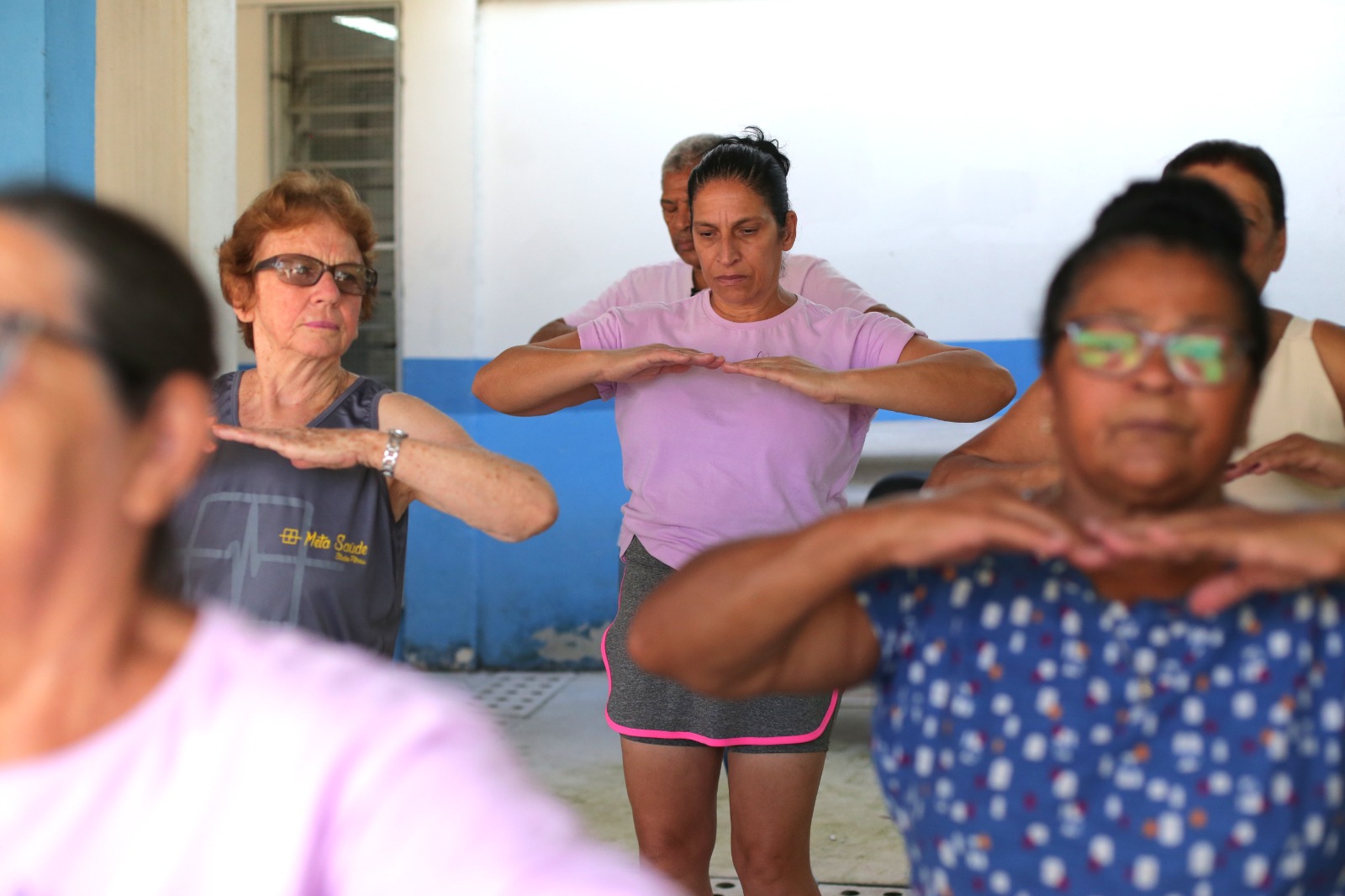 Foto mostra um grupo de adultos participando de uma atividade física coletiva em um equipamento da rede municipal de saúde. Em primeiro plano, uma mulher usa óculos e camiseta cinza, enquanto outra, ao centro, veste camiseta rosa e shorts cinza. Os participantes realizam um exercício com os braços elevados à altura do peito, acompanhando a orientação da atividade. Ao fundo, outras pessoas repetem os movimentos em um ambiente amplo e iluminado. A prática regular de exercícios físicos é uma das principais estratégias para o controle do colesterol e a prevenção de doenças cardiovasculares.