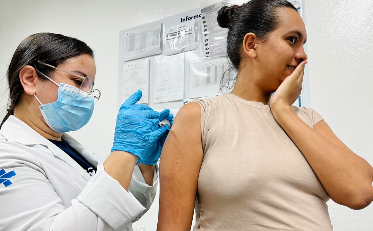 A imagem mostra uma profissional de saúde aplicando uma vacina no braço de uma mulher, em uma sala de vacinação. A profissional usa jaleco branco, máscara cirúrgica, óculos de proteção e luvas azuis, enquanto segura a seringa durante a aplicação. A paciente está com o braço exposto e leva uma das mãos ao rosto, em uma expressão que demonstra apreensão diante da vacinação. Ao fundo, há cartazes e documentos informativos afixados na parede, caracterizando o ambiente de uma unidade de saúde.