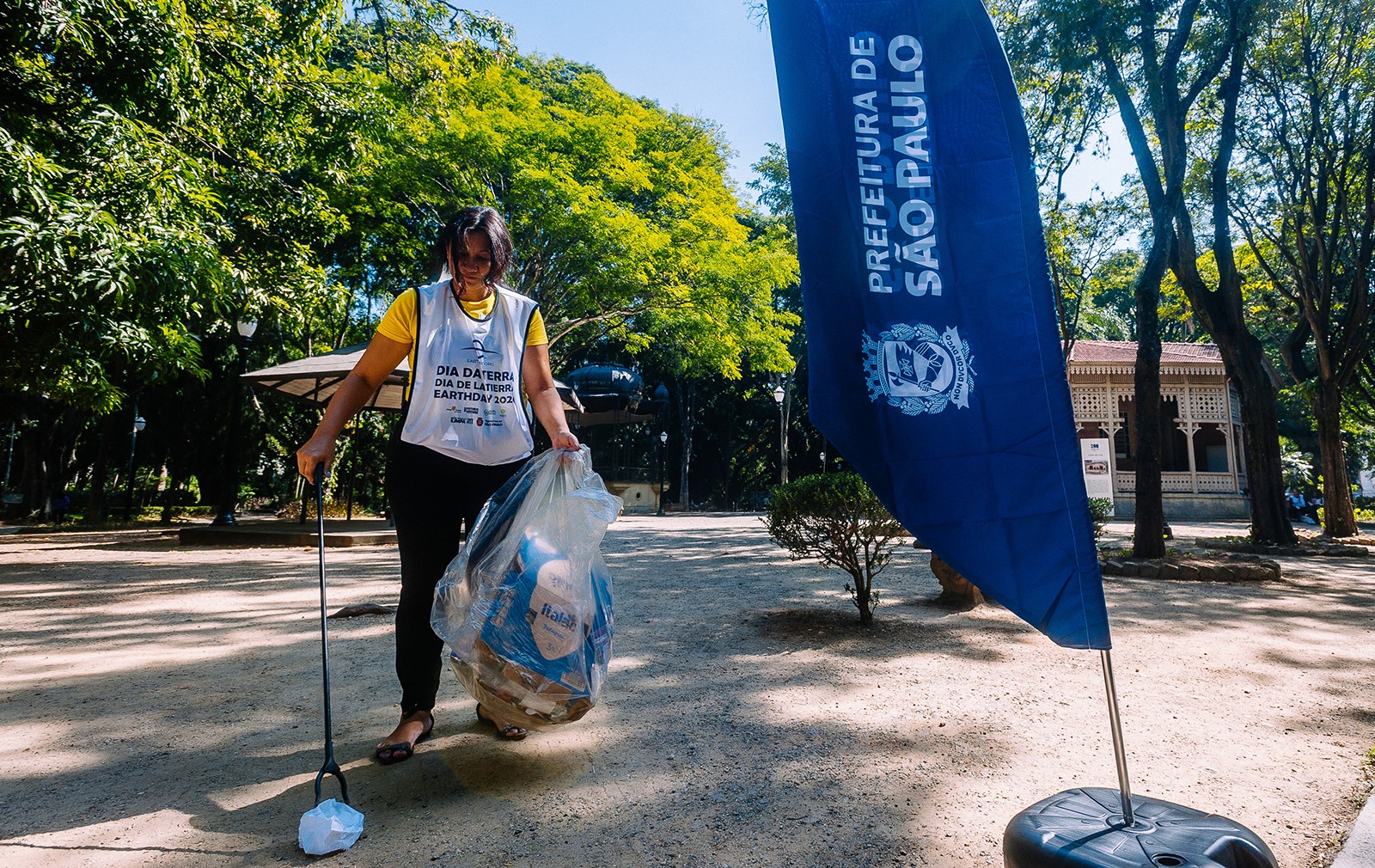 Em uma área arborizada de parque, uma mulher participa de uma ação de limpeza urbana. Ela veste camiseta amarela e colete branco com dizeres sobre o Dia da Terra. Com uma pinça longa na mão esquerda, recolhe um papel amassado do chão, enquanto segura um saco plástico transparente cheio de resíduos recicláveis na outra mão. Ao lado dela, há uma bandeira azul vertical com a inscrição “Prefeitura de São Paulo”. Ao fundo, aparecem árvores altas, caminhos de terra clara e uma construção antiga parcialmente visível entre a vegetação. O dia está ensolarado, com sombras das árvores espalhadas pelo chão.