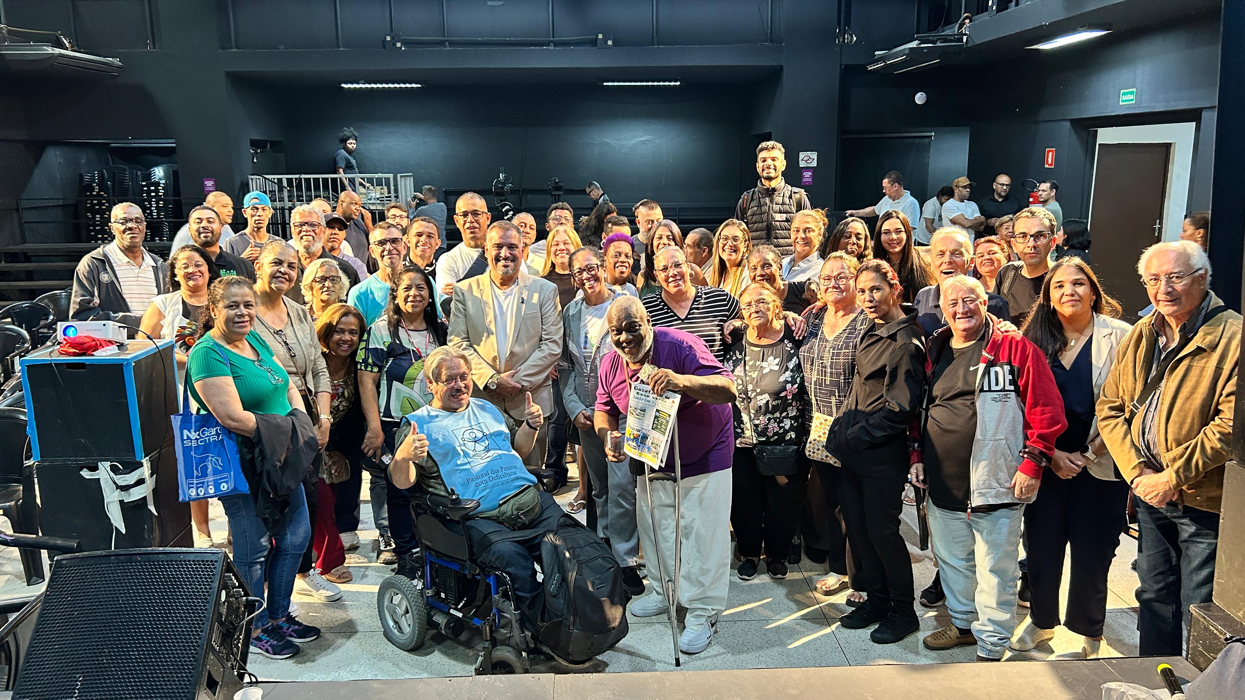 Foto realizada após o evento A imagem mostra um grande grupo diverso de cerca de 50-60 pessoas reunidas em um espaço interno, como um auditório o. Elas  posam  para uma foto em grupo, com destaque para um homem em cadeira de rodas no centro à esquerda fazendo sinal de positivo com as mãos.