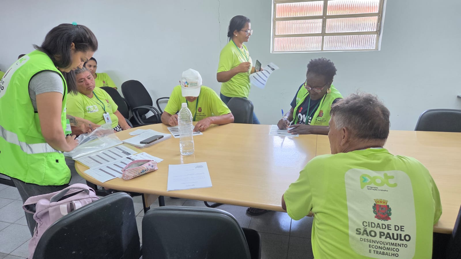 A imagem mostra um grupo de pessoas reunidas em uma sala simples, sentadas ao redor de uma mesa oval de madeira clara. A maioria veste camisetas verdes com identificação do POT.  À esquerda, uma mulher em pé, também de verde, está inclinada sobre a mesa, organizando ou entregando materiais dentro de um saco plástico. Ao redor da mesa, outras pessoas — homens e mulheres, em sua maioria adultos e idosos — estão sentadas, concentradas em preencher formulários com caneta.  