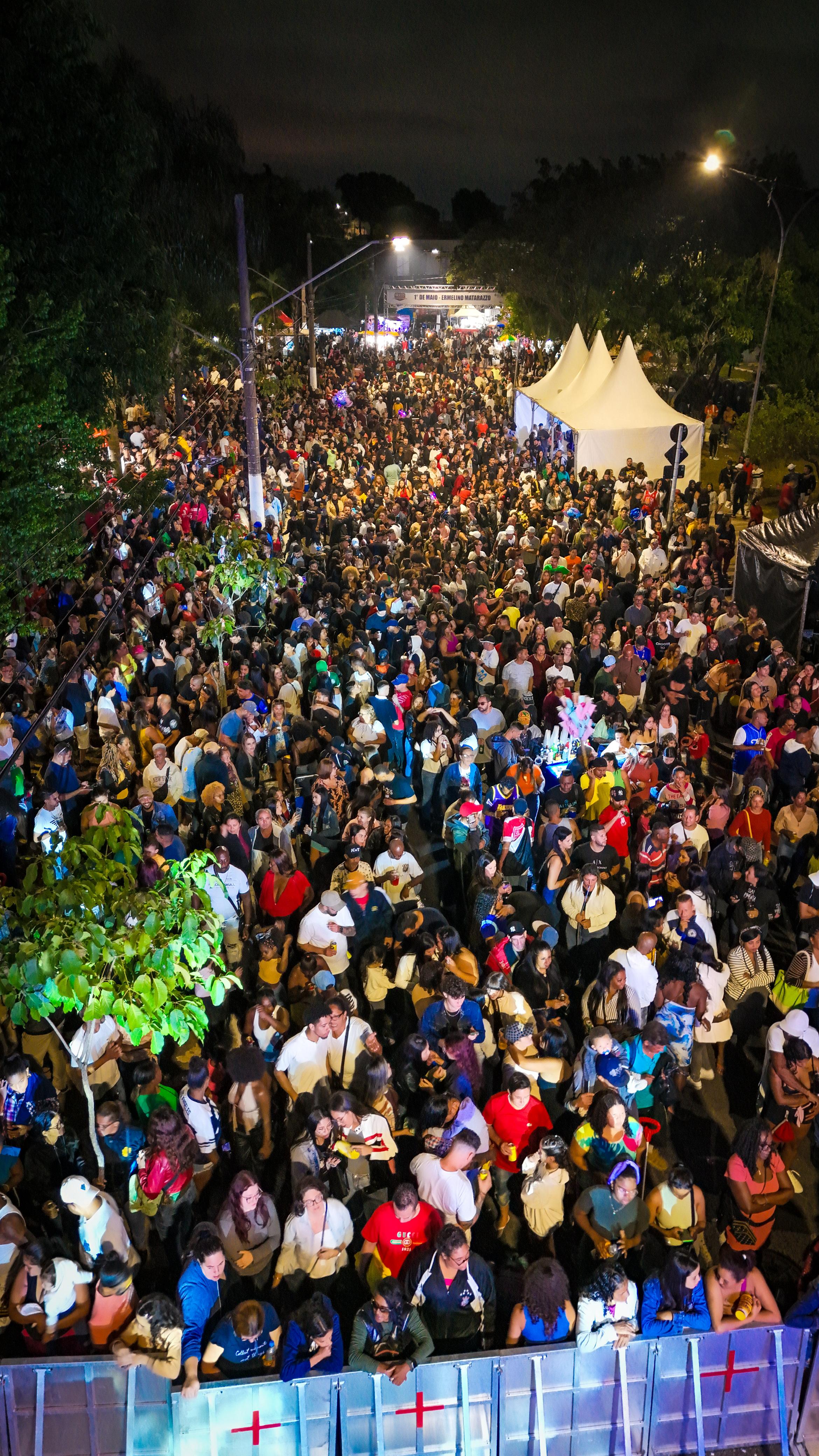 Uma fotografia de ângulo alto, tirada de um ponto de vista elevado (provavelmente do palco), mostrando uma enorme e densa multidão de pessoas reunida em uma rua durante a noite.  No canto inferior da imagem, em primeiro plano, aparece uma grade metálica de proteção cinza com detalhes vermelhos, onde as primeiras fileiras de pessoas estão encostadas, olhando em direção à frente. A multidão é diversa e se estende por toda a largura e comprimento da rua, sumindo ao fundo sob a iluminação noturna.  Do lado direito da rua, há grandes tendas brancas pontiagudas e, mais à frente, estruturas pretas de apoio com a palavra "SEGURANÇA" escrita em letras brancas. Do lado esquerdo, árvores de copas verdes ladeiam a via. A cena é iluminada por postes de luz amarelada e por um forte refletor ao fundo, que projeta um feixe de luz brilhante e azulada sobre o público. O céu está escuro.
