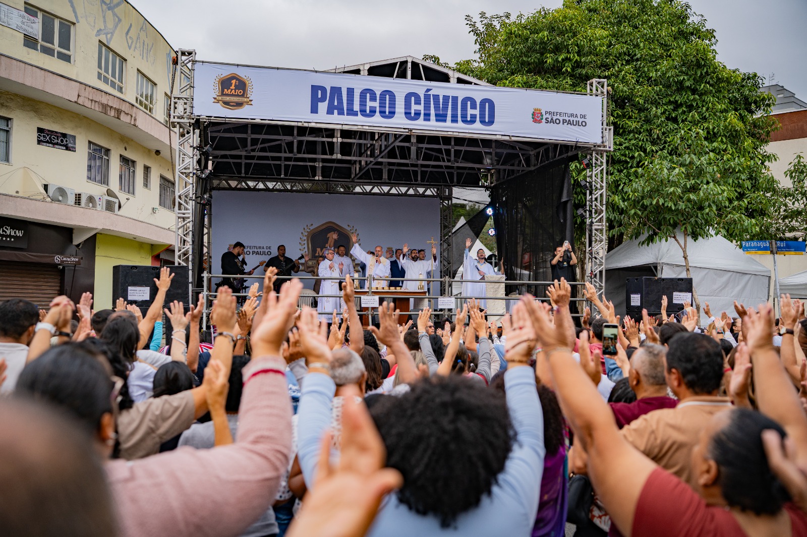 Uma fotografia de média-distância tirada por trás de uma multidão em pé na rua, com os braços erguidos em direção a um palco coberto ao ar livre, onde um homem de branco comanda a cerimônia com outros homens no palco. O palco é uma estrutura metálica com uma lona branca esticada no topo, com "PALCO CÍVICO" escrito em azul no centro da lona. À direita da escrita está o brasão da Prefeitura de São Paulo e abaixo dele, as palavras "PREFEITURA DE SÃO PAULO". No palco, o homem está de pé diante de um altar de madeira com itens de metal dourado e outros itens sobre ele, e os outros homens estão de pé atrás dele. Um dos homens de pé atrás do homem de branco está com os braços erguidos para o alto. Fora do palco, no canto inferior direito da imagem, uma placa de rua azul e branca que lê "Avenida São João" e, abaixo dela, "Rua" está em pé sobre um poste, inclinada para a direita da imagem, enquanto uma tenda branca está atrás da placa. No canto inferior esquerdo da imagem está uma placa de rua azul e branca com "Show" escrito em branco e uma flecha branca apontando para a direita da imagem.