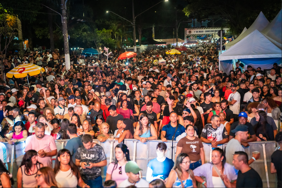 Uma fotografia colorida em plano médio e ângulo ligeiramente alto, mostrando uma grande e compacta multidão de pessoas reunida em uma rua durante um evento noturno.  Na metade inferior da imagem, uma grade de metal cinza serve como barreira de segurança, separando o público do primeiro plano. Atrás da grade, centenas de pessoas de perfis diversos aparecem em pé, espremidas umas contra as outras, olhando predominantemente em direção à esquerda (onde estaria o palco). Entre o público, destacam-se alguns guarda-sóis abertos: um listrado de amarelo e branco no lado esquerdo e um vermelho ao fundo.  No canto superior direito, veem-se grandes tendas brancas com coberturas pontiagudas iluminadas por dentro. Ao fundo, no centro da imagem, há um pórtico ou portal iluminado que atravessa a rua, onde é possível ler parcialmente as palavras "1º DE MAIO - ERMELINO MATARAZZO". A iluminação pública artificial cria reflexos nos rostos das pessoas, e o topo do cenário exibe copas de árvores escuras contra o céu da noite.
