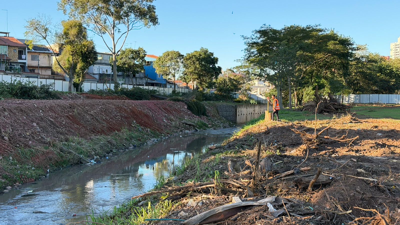 A imagem mostra um canal ou córrego de água turva cortando uma área urbana em obras, sob a luz clara do dia.  O canal corre do centro do fundo em direção ao primeiro plano, no canto inferior esquerdo. O reflexo do céu azul e de parte da vegetação pode ser visto na superfície da água, que contém alguns pequenos detritos flutuantes.  Detalhes das margens: Margem esquerda: É formada por um barranco inclinado de terra avermelhada e rasteira, sem vegetação densa. No topo desse barranco, há um muro branco que separa a área do canal de um bairro residencial com casas de dois ou três andares e várias árvores altas.  Margem direita: Apresenta uma área plana de terra batida coberta por muitos galhos secos, restos de troncos e raízes acumulados, evidenciando uma limpeza ou desmatamento recente. Mais ao fundo, o terreno se transforma em uma área gramada e arborizada.