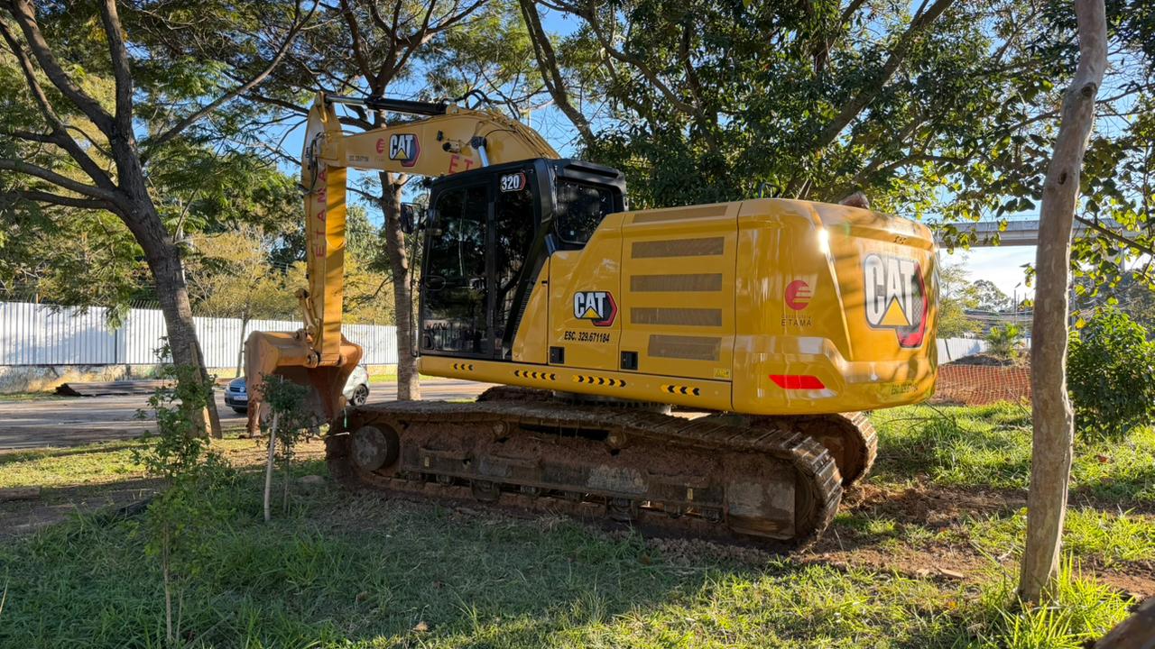 A imagem mostra uma escavadeira hidráulica de grande porte, da marca CAT (Caterpillar), estacionada em uma área gramada e arborizada sob a luz do dia.  O veículo é predominantemente amarelo, com detalhes em preto na cabine e nas esteiras de metal. Ele está posicionado em diagonal, com a parte traseira e a lateral direita mais visíveis em primeiro plano, enquanto o braço mecânico e a caçamba se estendem para o lado esquerdo, parcialmente ocultos pelas árvores.  Detalhes do veículo: Traseira e Lateral: Na parte traseira amarela, destaca-se o logotipo cinza e vermelho da "CAT". Na lateral, há a inscrição "CAT" em um fundo preto, acompanhada do texto menor "ESC. 329.671184". Mais acima, próximo à cabine preta, vê-se o número "320".  Esteiras: As esteiras de tração, feitas de metal escuro, estão apoiadas diretamente sobre a terra batida e a grama.  Cenário ao fundo: A escavadeira está cercada por árvores de copas verdes que projetam sombras pelo chão.  Ao fundo, no lado esquerdo, há uma cerca ou muro branco texturizado e um carro de cor escura estacionado na rua.  O céu aparece azul e limpo entre as folhas das árvores, indicando um dia ensolarado.