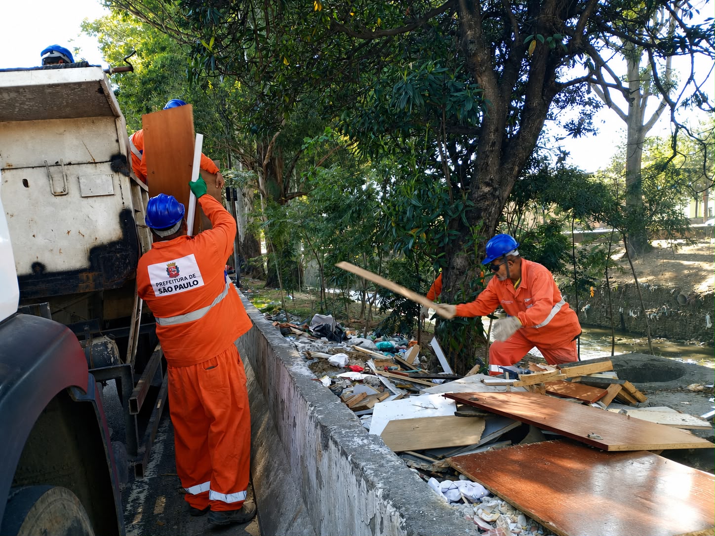 Foto em plano médio e ângulo normal, tirada ao ar livre durante o dia, sob luz solar direta.  Três trabalhadores vestindo uniformes de sinalização na cor laranja, com faixas refletivas e capacetes azuis, realizam a limpeza de entulhos e resíduos ao lado de uma mureta de concreto.  Em primeiro plano, à esquerda, as costas de um caminhão basculante branco está estacionado. Um dos trabalhadores, visto de costas, ergue uma grande prancha de madeira em direção à caçamba do caminhão; na parte posterior de seu uniforme, há o logotipo e a inscrição "PREFEITURA DE SÃO PAULO".  À direita, outro trabalhador, usando óculos escuros e luvas brancas, está agachado ao lado de uma pilha de pedaços de madeira, compensados e resíduos, arremessando uma ripa de madeira para cima. Ao fundo, há árvores de copa densa, um canal com vegetação e um trecho de terreno ensolarado.