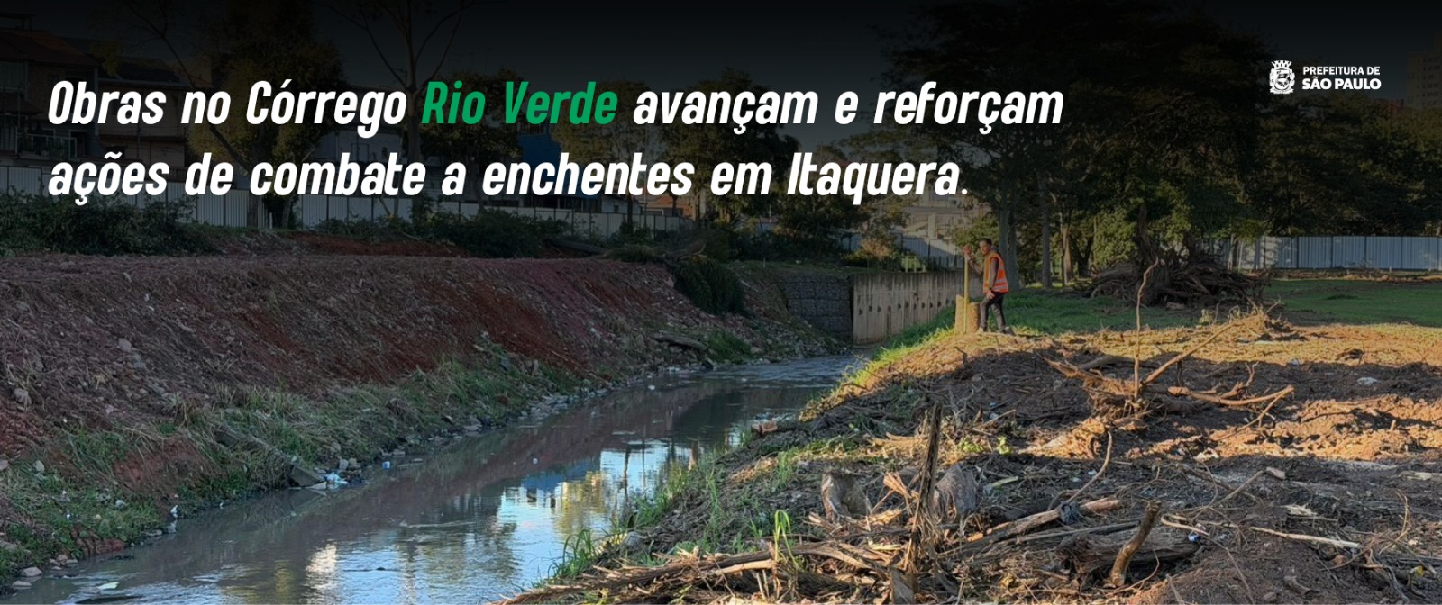 A imagem mostra uma foto panorâmica diurna de uma obra de canalização e contenção de enchentes em um córrego urbano.  No centro, o Córrego Rio Verde corre em um canal estreito com água de tom cinzento. A margem esquerda do córrego é um barranco inclinado de terra avermelhada misturada com um pouco de vegetação rasteira. A margem direita exibe uma área de terra batida revirada, com galhos secos, raízes e restos de vegetação acumulados em primeiro plano. Mais ao fundo, na mesma margem, há um gramado verde com árvores e um trabalhador vestindo um colete refletivo alaranjado em pé, observando o local. Ao fundo, uma barreira de concreto protege parte da encosta do canal.  Na parte superior da imagem, há uma faixa escura translúcida onde se destaca um texto em letras brancas e verdes com inclinação itálica. No canto superior direito, vê-se o logotipo oficial da Prefeitura de São Paulo.
