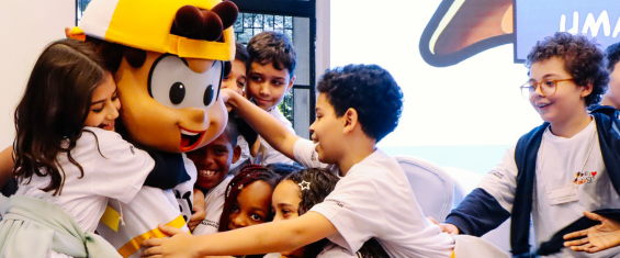 Fotografia colorida em ambiente interno mostrando um grupo de crianças reunidas em torno de um personagem mascote de desenho animado. O personagem usa boné amarelo, camiseta branca e calça azul, e está sentado enquanto recebe um abraço coletivo das crianças. Meninos e meninas, vestindo camisetas brancas, sorriem e interagem com entusiasmo, demonstrando carinho e alegria. Ao fundo, há uma grande janela que permite a entrada de luz natural e um painel com ilustração colorida. O clima da cena é de diversão, acolhimento e participação infantil em uma atividade educativa ou recreativa.