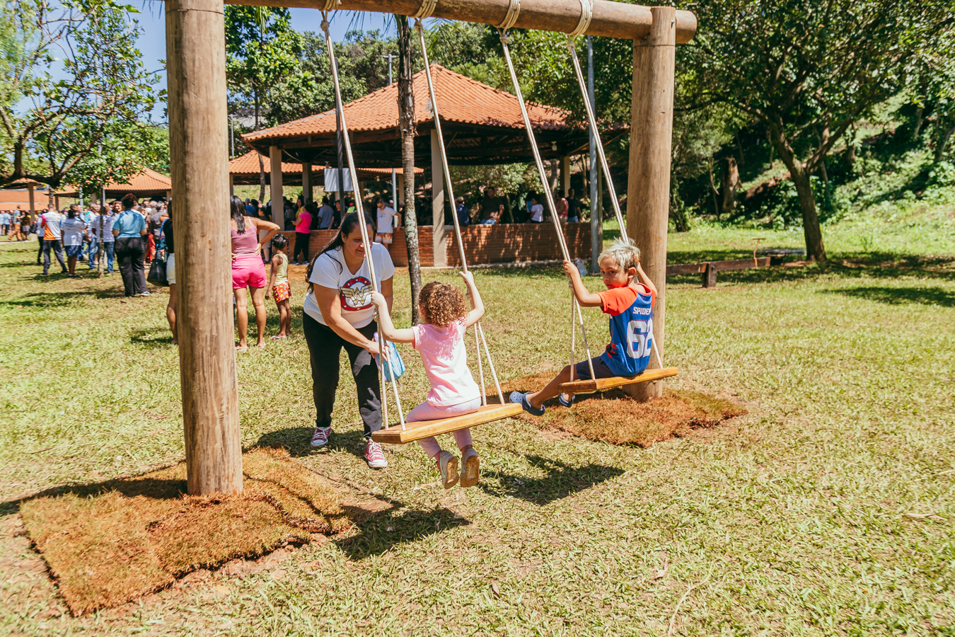 Em um parque ensolarado, duas crianças brincam em um balanço de madeira instalado sobre um gramado. Uma mulher empurra delicadamente uma menina de cabelos cacheados, enquanto um menino, sentado no balanço ao lado, olha em direção à câmera. Ao fundo, diversas pessoas estão reunidas próximas a um quiosque de telhado cerâmico, cercado por árvores de grande porte que compõem a paisagem verde do parque. A cena transmite um momento de lazer, convivência e contato com a natureza.