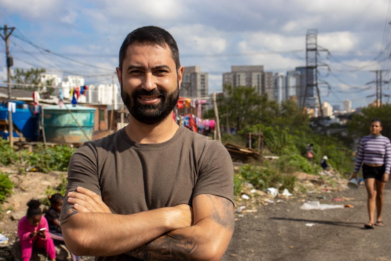 Uma imagem em plano médio mostra um homem de pele clara, com barba e cabelos curtos e escuros, sorrindo para a câmera com os braços cruzados. Ele veste uma camiseta marrom e possui tatuagens visíveis nos antebraços.  O homem está posicionado em primeiro plano, no centro da imagem. Ao fundo, à esquerda, há uma área residencial simples com construções de tijolos aparentes, varais com roupas coloridas e uma caixa d'água azul. No canto inferior esquerdo, duas crianças estão sentadas no chão.  À direita, uma mulher caminha por uma estrada de terra. Ao fundo, a paisagem urbana revela prédios altos sob um céu azul com nuvens brancas, além de torres de transmissão de energia elétrica. A iluminação é natural e frontal, destacando o rosto do homem.