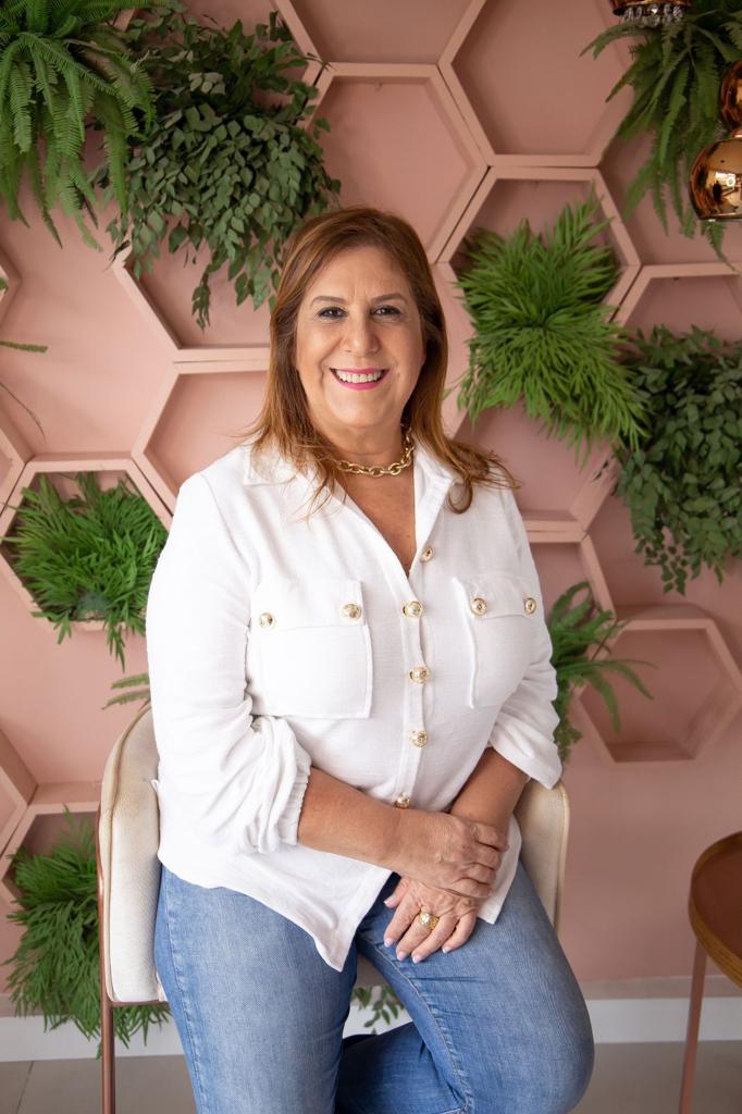 Foto da secretária municipal da Pessoa com Deficiência, Silvia Grecco. Mulher sentada em uma cadeira, posando para um retrato. Ela tem pele clara, cabelos castanhos, de comprimento médio, na altura dos ombros, e está sorrindo, olhando para a frente. Usa uma camisa social branca, com botões dourados grandes e bolsos na altura do peito, mangas dobradas e calça jeans azul. No pescoço, usa um colar curto dourado e, nas mãos, alguns anéis. As mãos estão apoiadas uma sobre a outra, sobre a perna. O fundo é uma parede rosa-clara, preenchida, em vários pontos, por folhagens verdes.