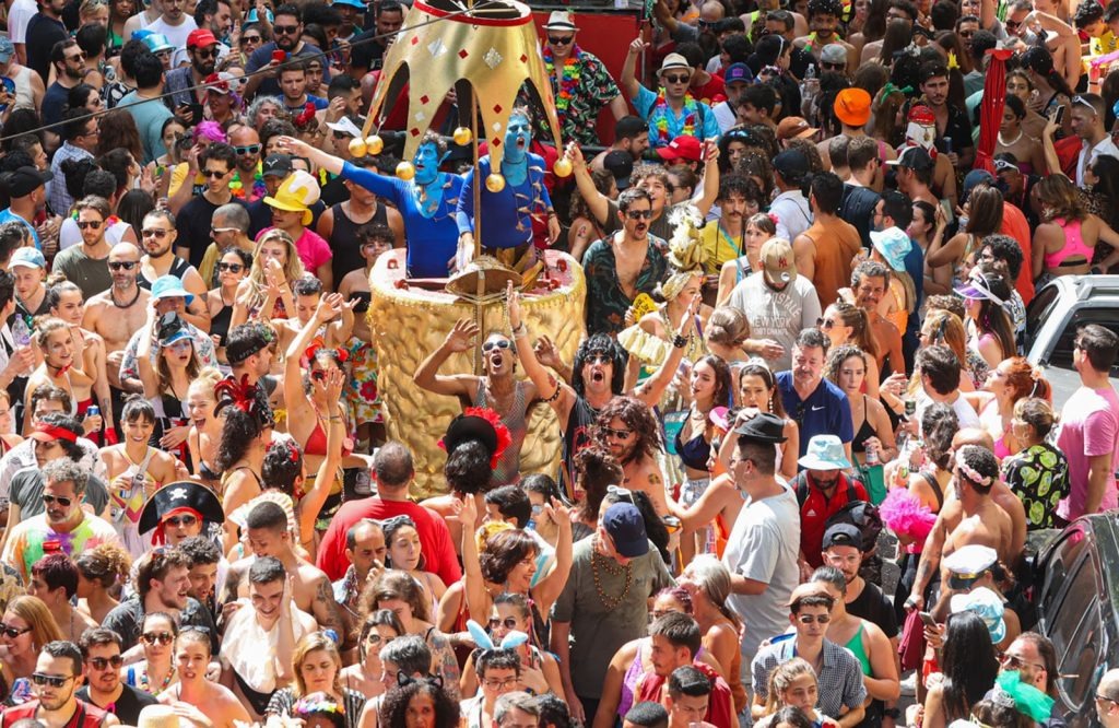 Foto em plano geral de uma multidão densa e animada em um bloco de carnaval de rua. No centro, há uma estrutura dourada e ornamentada, semelhante a um carro alegórico, onde estão duas pessoas pintadas de azul, com adereços na cabeça, acenando. Ao redor, centenas de foliões de todas as idades e raças celebram, vestindo roupas coloridas, fantasias diversas, chapéus e adereços. Muitos sorriem, dançam e levantam os braços. A luz do dia é natural e forte, iluminando a cena. A rua está completamente tomada pela festa, transmitindo uma sensação de alegria e celebração.