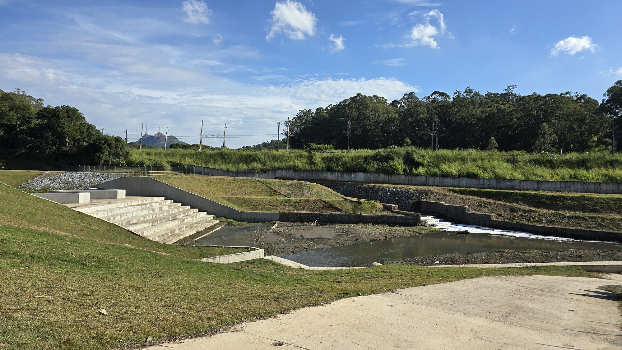 Imagem mostra uma área aberta com estrutura de drenagem e contenção de água cercada por vegetação. Em primeiro plano, há um reservatório com pequeno fluxo de água, escadaria de concreto e taludes gramados. Ao fundo, aparecem árvores, postes de energia e trilhos ferroviários sob céu azul com nuvens dispersas.