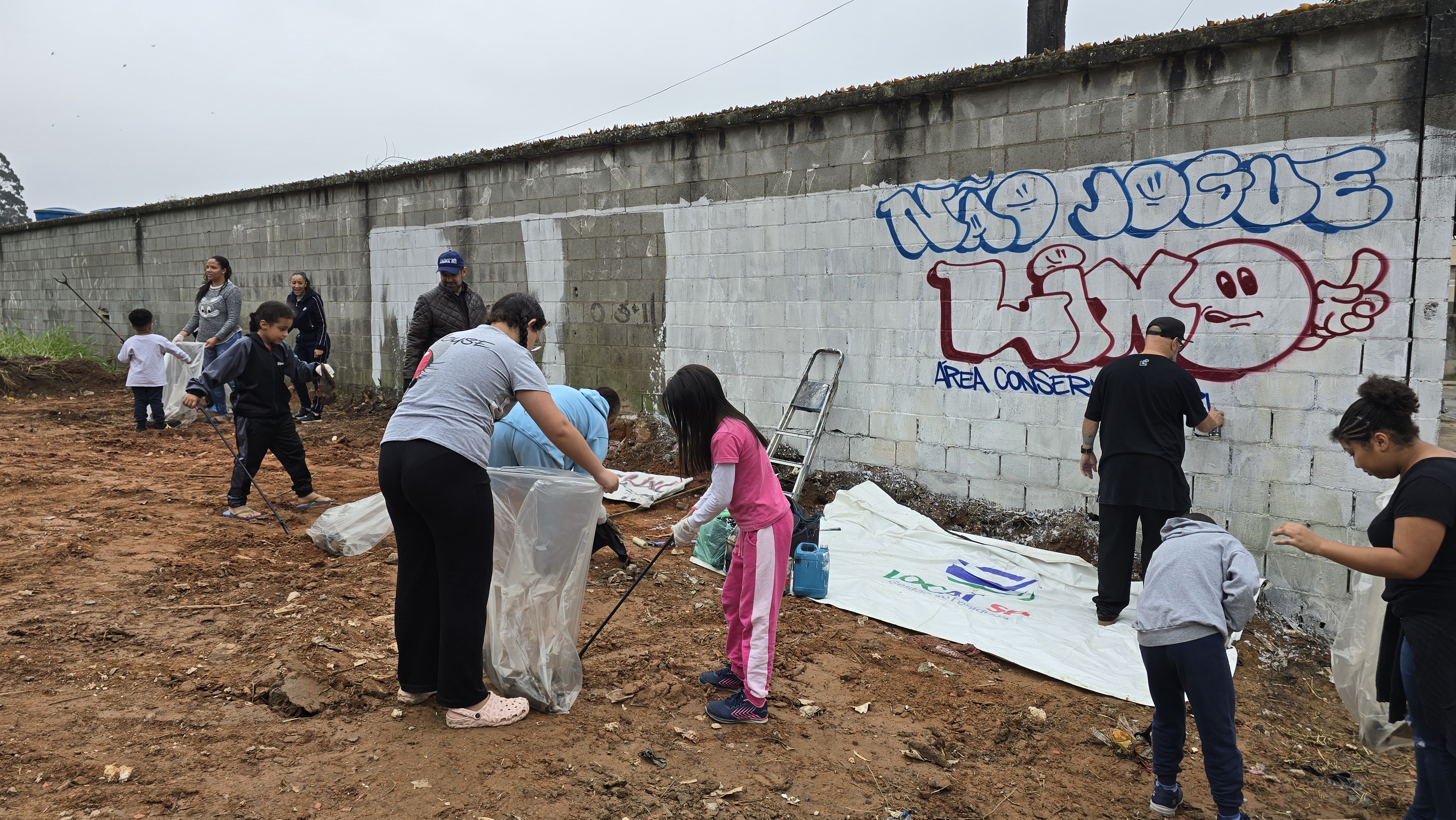 A imagem mostra um mutirão comunitário de revitalização urbana realizado em uma área aberta de solo exposto. Em primeiro plano, moradores de diferentes idades, incluindo crianças e adultos, participam da limpeza do local utilizando sacos plásticos e ferramentas de coleta de resíduos.  Ao fundo, há um longo muro de blocos de concreto parcialmente pintado de branco, onde duas pessoas trabalham na cobertura de pichações. Em uma das extremidades do muro ainda é possível ver um grafite em azul e vermelho com a frase "Área Conserv..." parcialmente visível.  O ambiente apresenta aspecto de transformação e cuidado coletivo, com voluntários distribuídos pelo terreno em diversas atividades simultâneas. O céu está nublado, e a cena transmite a ideia de mobilização social, preservação do espaço público e recuperação ambiental.