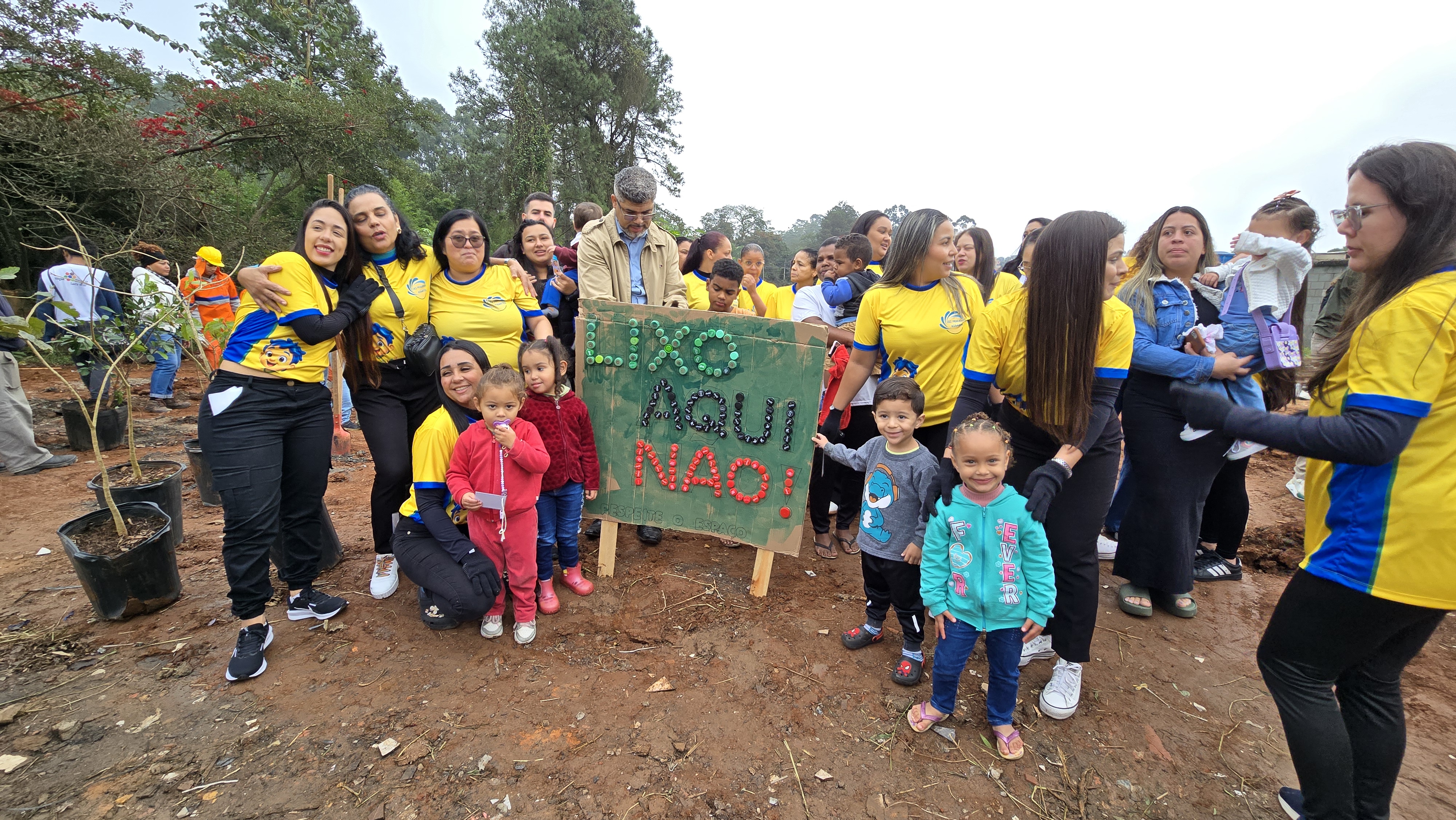 Imagem mostra um grupo de adultos e crianças participando de uma ação de plantio coletivo em uma área aberta. Os participantes, muitos usando camisetas amarelas, posam ao redor de uma placa com a mensagem “Fixe aqui não!”, enquanto ao lado há mudas de árvores prontas para o plantio. Ao fundo, outras pessoas realizam atividades de arborização, reforçando o caráter comunitário e ambiental da iniciativa.