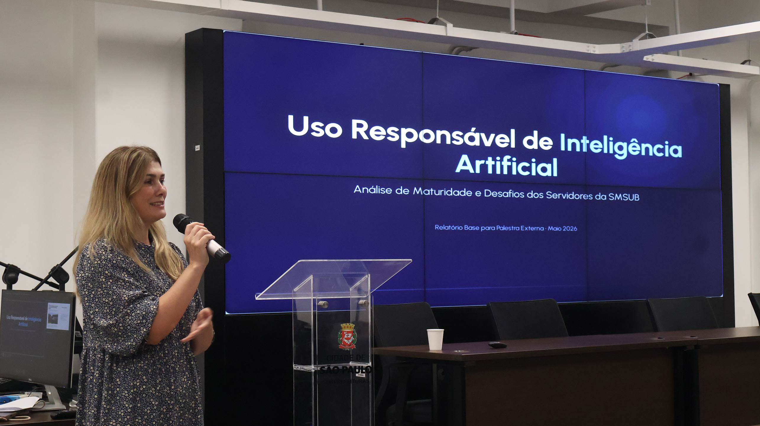 Imagem mostra uma apresentação realizada em auditório da Prefeitura de São Paulo sobre o tema “Uso Responsável de Inteligência Artificial”. Uma mulher segura um microfone enquanto fala ao público ao lado de um púlpito com o brasão da cidade. Ao fundo, um grande telão exibe o título da palestra e informações sobre análise de maturidade e desafios relacionados ao uso de IA na SMSUB.