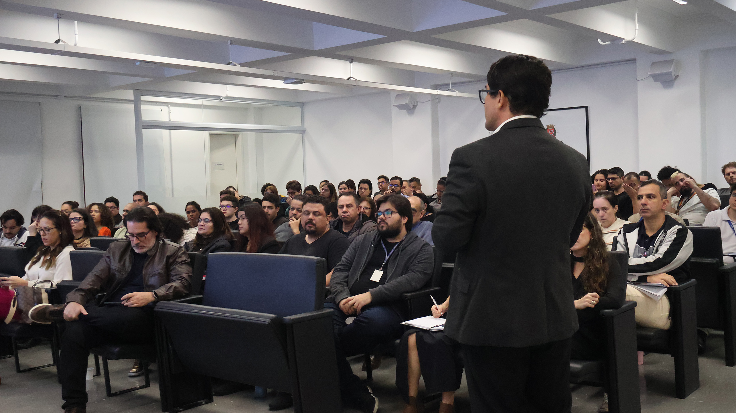 Imagem mostra uma palestra ou reunião realizada em um auditório da Prefeitura de São Paulo. Um homem de terno aparece em primeiro plano falando para o público, enquanto dezenas de pessoas acompanham a apresentação sentadas em poltronas. O ambiente é fechado, com iluminação interna e participantes atentos à atividade.