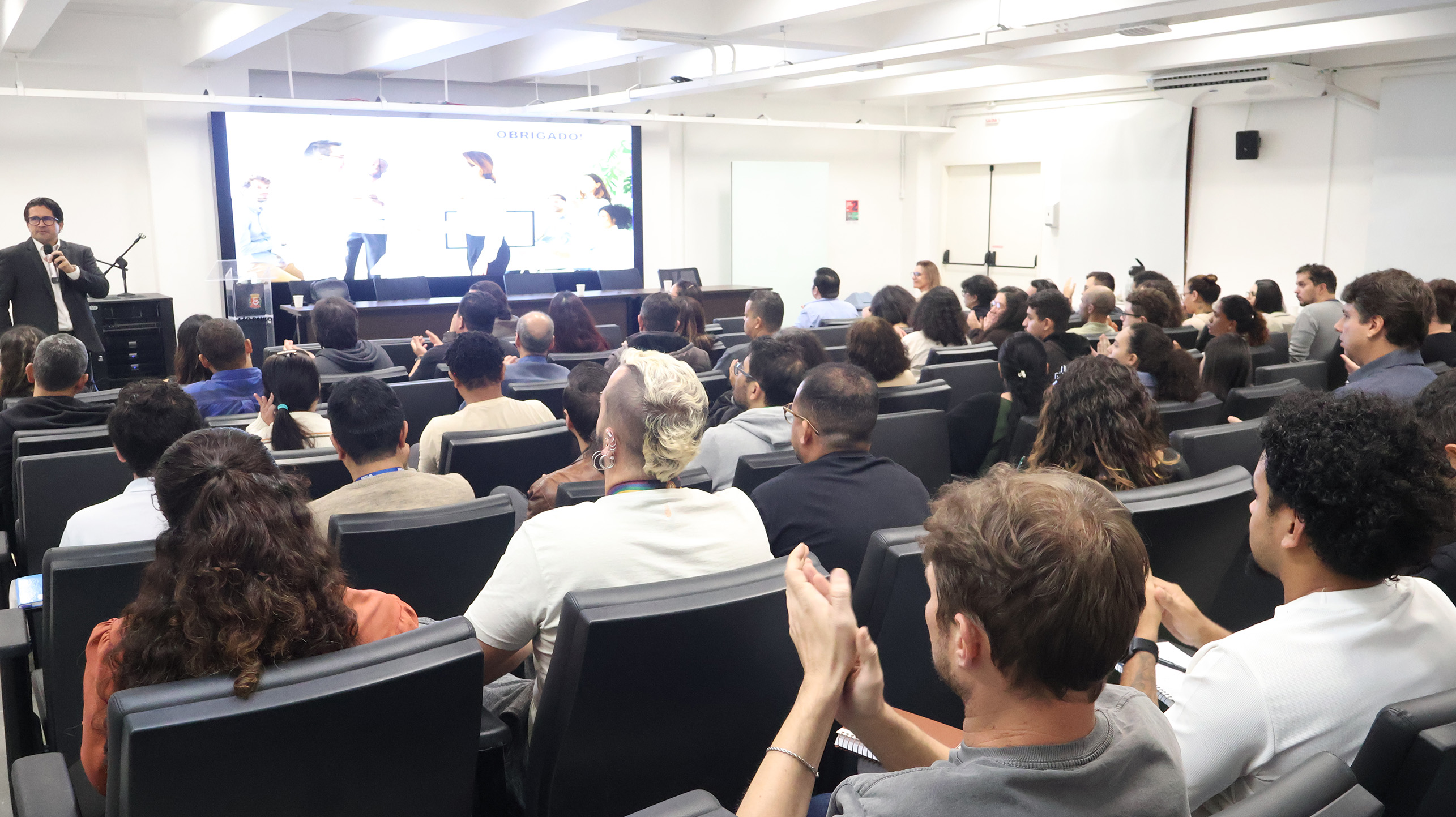 Imagem mostra um auditório lotado durante uma palestra ou evento promovido pela Prefeitura de São Paulo. Os participantes estão sentados em poltronas acompanhando uma apresentação exibida em um grande telão ao fundo. À esquerda, um homem fala ao público com microfone na mão, enquanto parte da plateia aparece aplaudindo ao final da atividade.