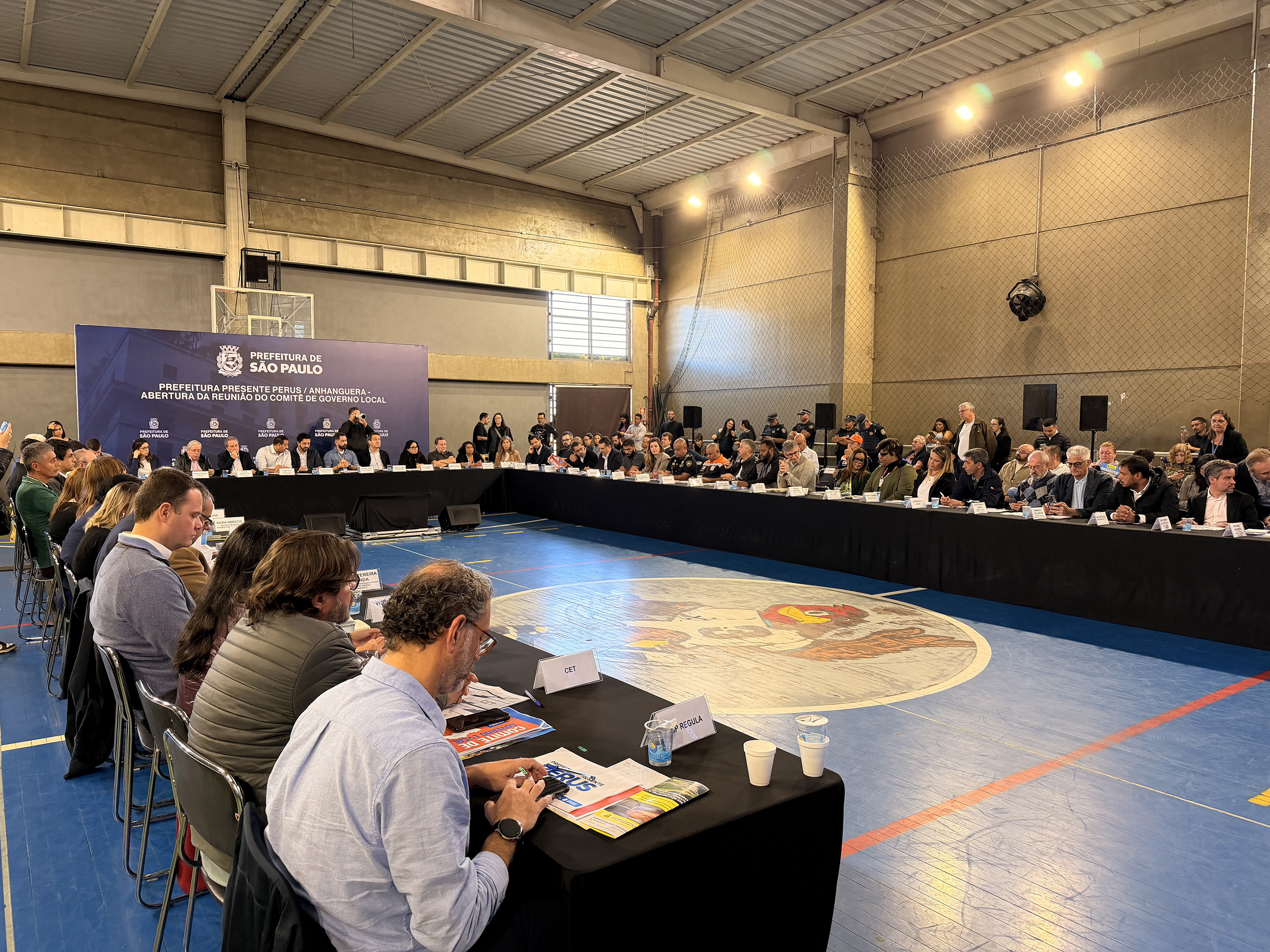 Imagem mostra uma reunião institucional promovida pela Prefeitura de São Paulo em um ginásio esportivo. Autoridades, representantes de órgãos públicos e participantes estão sentados ao redor de uma grande mesa em formato de “U”. Ao fundo, um painel informa a abertura da reunião do Comitê de Crise na região de Perus e Anhanguera. O espaço possui iluminação interna ampla e quadra esportiva ao centro.