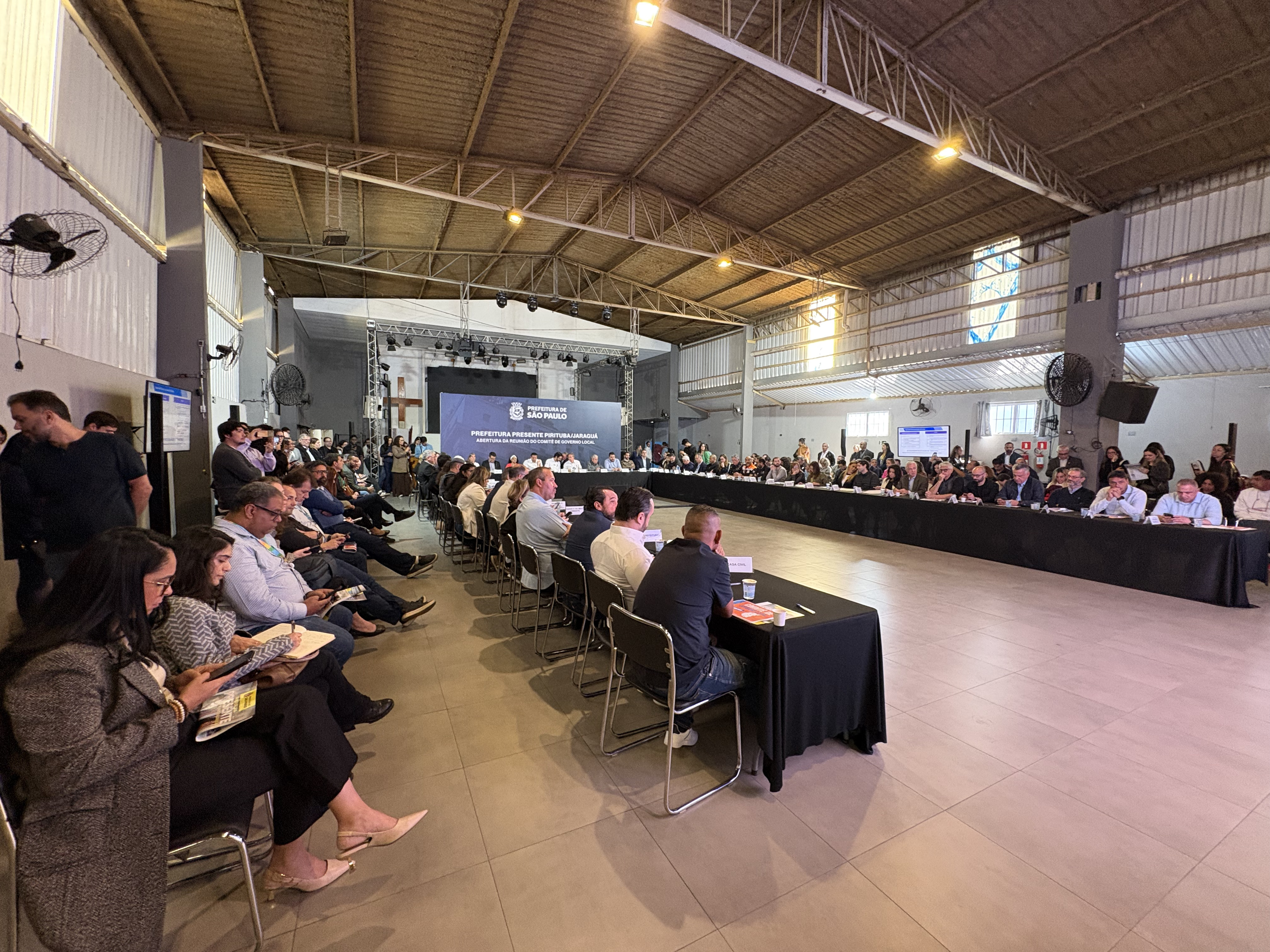 Foto de um grande salão industrial, com teto alto de estrutura metálica e iluminação quente. O espaço está lotado para um evento da Prefeitura de São Paulo. Ao fundo, há um palco com um telão exibindo a identidade visual da Prefeitura e informações sobre uma reunião. Em frente ao palco, dezenas de pessoas estão sentadas em mesas organizadas em formato de “U”, acompanhando a atividade. Outras pessoas estão sentadas em fileiras nas laterais. No centro e nas laterais do salão há um amplo espaço livre. A imagem mostra um ambiente formal, com autoridades, servidores e participantes reunidos para uma apresentação ou reunião institucional.