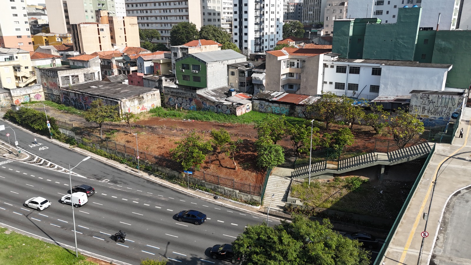 Imagem aérea mostra uma área urbana próxima a uma avenida movimentada na cidade de São Paulo. O espaço possui terreno aberto com árvores, vegetação e muros com grafites ao redor. À direita, há uma passagem elevada para pedestres sobre um córrego canalizado. Ao fundo, aparecem casas, prédios residenciais e comerciais característicos da paisagem urbana da capital paulista.