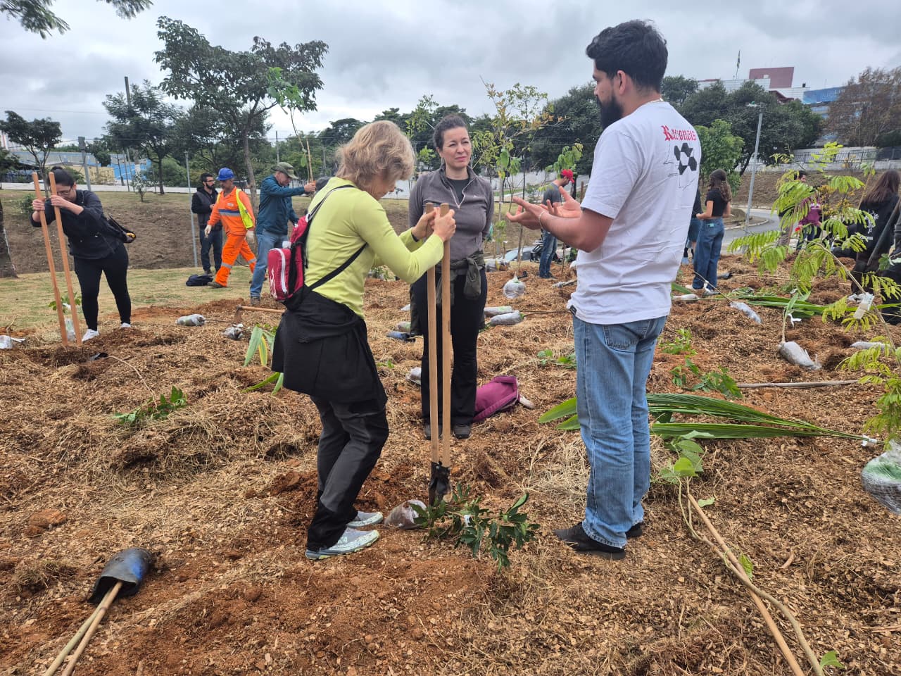 Imagem mostra um mutirão de plantio de árvores realizado em uma área aberta e recém-preparada para recuperação ambiental. Voluntários e participantes utilizam ferramentas de jardinagem para plantar mudas nativas, enquanto outras pessoas auxiliam na organização e no manejo do terreno. Ao fundo, é possível ver novas mudas já plantadas, áreas cobertas com matéria orgânica e vegetação, sob céu nublado.